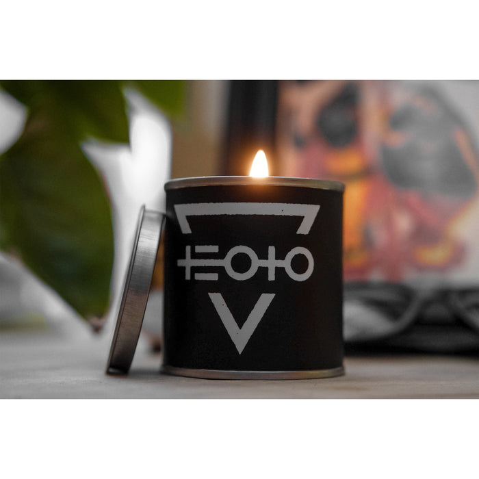 A black tin candle from The End Of The Ocean, with a white geometric pattern of lines and circles, is lit with its lid nearby. A green plant and a blurred background add to the calm ambiance.
