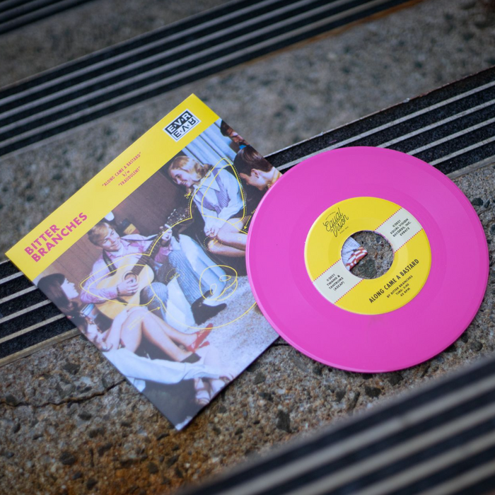 Vinyl jacket with an image of people sitting in a circle and watching a boy play guitar. There is a pig faintly drawn in yellow. There is a yellow ribbon across the top. On the top left, "BITTER BRANCHES" is written in red lettering. Towards the center, "ALONG CAME A BASTARD" is written in smaller red lettering. To the top right, there is a rectangle with EVR written on top and bottom. It is written normally on top, but it is being mirrored on bottom. There is a pink disc laying on concrete stai