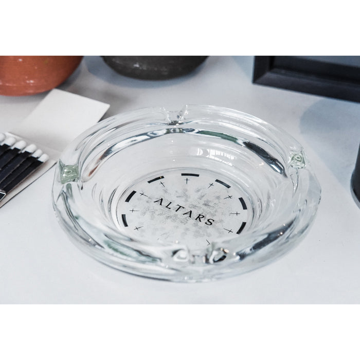 A clear glass Ash Tray from Sadistik with “ALTARS” printed in the center rests on a white surface, alongside a box of matches and other items.