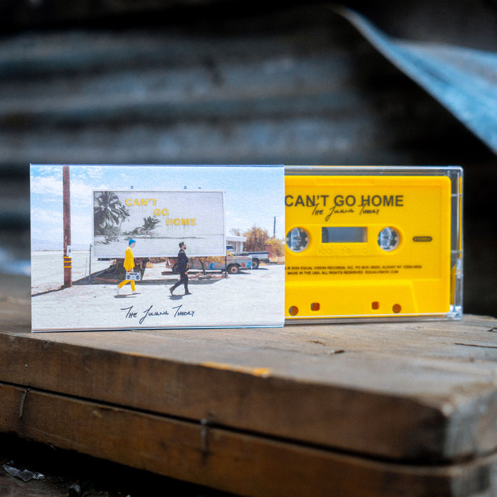 Cassette case with two people walking away from a beach, in front of a sign that says CAN'T GO HOME. In the background are some trucks behind the sign, and to the left is a pole. In the bottom center is black text that says THE JULIANA THEORY. Peeking out of the case is a yellow cassette that says CAN'T GO HOME on the top, and THE JULIANA THEORY below it. Both are propped up on a wooden plank.