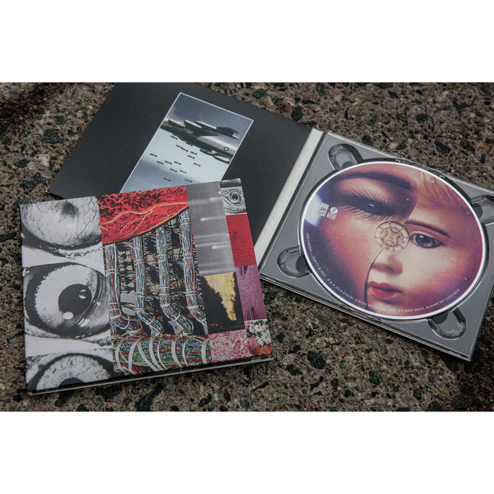 Square CD with a collage of images overlapping each other. Some of the images are black and white photos of eyes, and the others are various patterns. In the center there is a white rectangle with GREYHAVEN written in it. Below that is text that says EMPTY BLACK. Inside the CD cover is the disc, which is half of a doll's face and half of a girl's face. Both are laying on an asphalt surface.