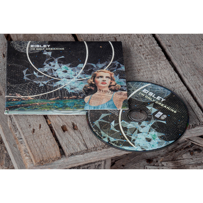 Square CD with album art of a woman looking off into the distance. In the background there is a loop design drawn in white and scenery that appears to be a mix of outer space and and mountains on earth. In the top left corner there is text that says EISLEY I'M ONLY DREAMING. Peeking out of the side is a CD with the same text on it and partial album cover design on it. Both are laying on top of a wooden surface.