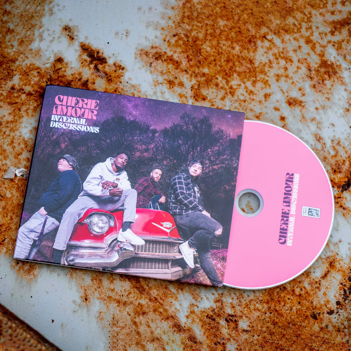 Square CD with an image of band members sitting on top of a red car. In the background are trees and the night sky, which is tinted pink along with the rest of the cover. In the top left corner there is pink text that says CHERIE AMOUR. Below that is white text that says INTERNAL DISCUSSIONS. To the right of the cover is a pink disc with CHERIE AMOUR written in purple and INTERNAL DISCUSSIONS written below in white. both are laying on top of an orange and white rusted surface.