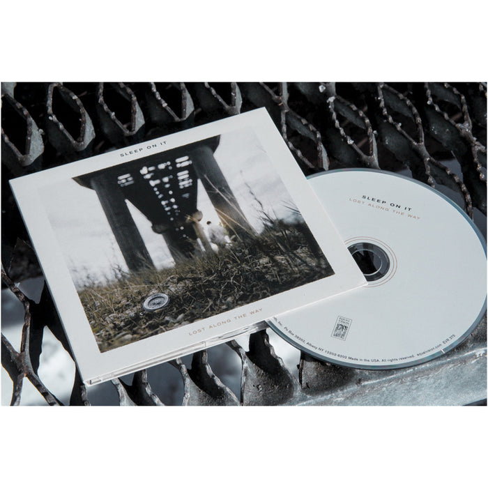 A Sleep On It CD and its case sit on a metal grate. The cover art shows a person under a sunlit bridge. The case is labeled Sleep On It - Lost Along The Way EP, with the track list featuring Counting Miles.