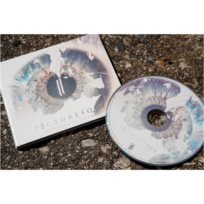Faded white CD case with a human eye faded onto the background. There is text at the bottom that reads Picturesque and Monstrous Things. Next to the cover is a cd all of which is laying on top of a road.