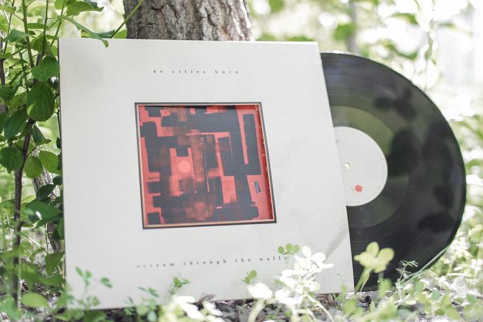 A vinyl record and cover by As Cities Burn, featuring abstract red artwork and “Scream Through The Walls” text, rest outdoors against a tree amid lush green plants. This is a limited pressing release.