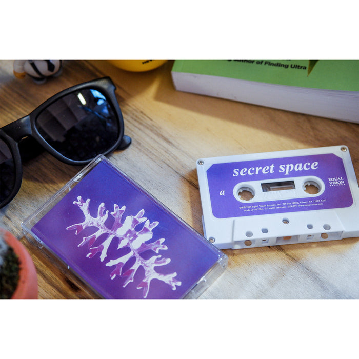 A white Secret Space cassette with a purple abstract-art case rests on a wooden surface beside black sunglasses and a green book.