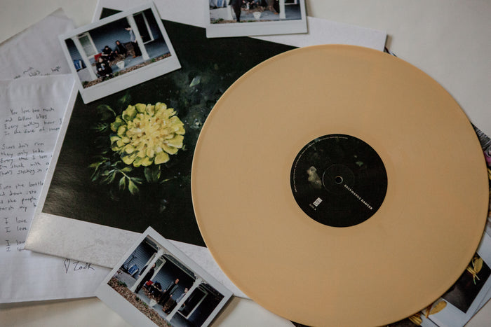 A beige Backwards Dancer self-titled vinyl rests on its album cover, displaying a yellow flower. Handwritten notes and instant photos of people outside a house surround the in-stock Backwards Dancer LP.