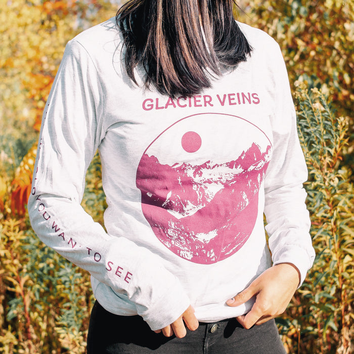 Athletic grey colored long sleeve shirt with GLACIER VEINS written in purple font across the chest. There is purple text down one of the sleeves that says THE WORLD YOU WANT TO SEE. On the cheset, there is a circular drawing of mountains and a moon inside it. An individual is modeling the shirt and standing in front of tall bushes.
