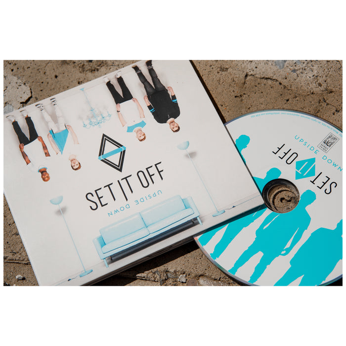 A Set It Off Upside Down album cover and CD are displayed on a stone surface. The cover shows five people upside down over a light blue couch, with Set It Off and Upside Down in bold lettering beside the track list.