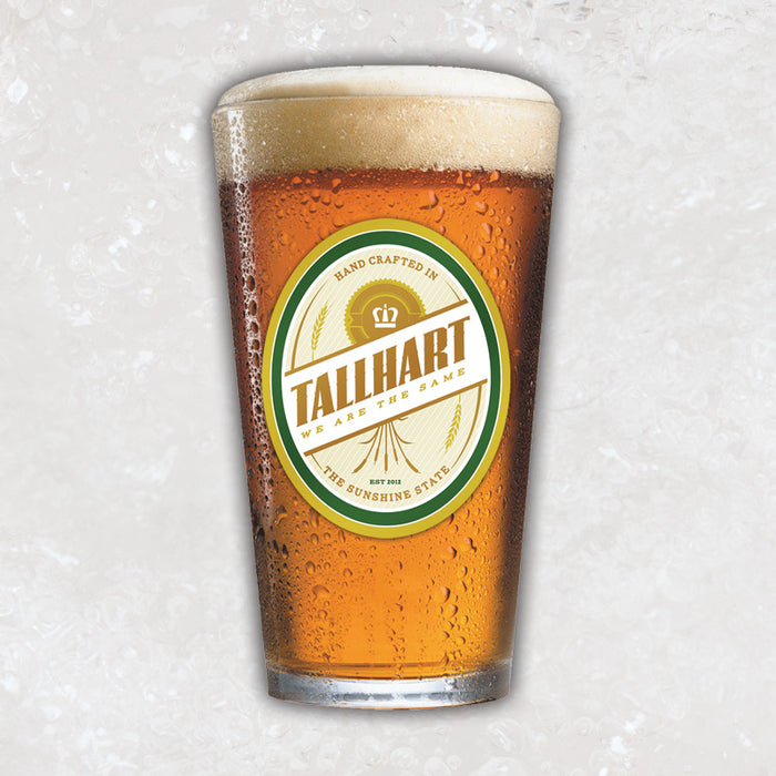 A pint glass with a oval shaped oat colored emblem. On the emblem it says "TALLHART" in diagonal lettering. There is also text on the top and bottom that says "HAND CRAFTED IN" "THE SUNSHINE STATE".