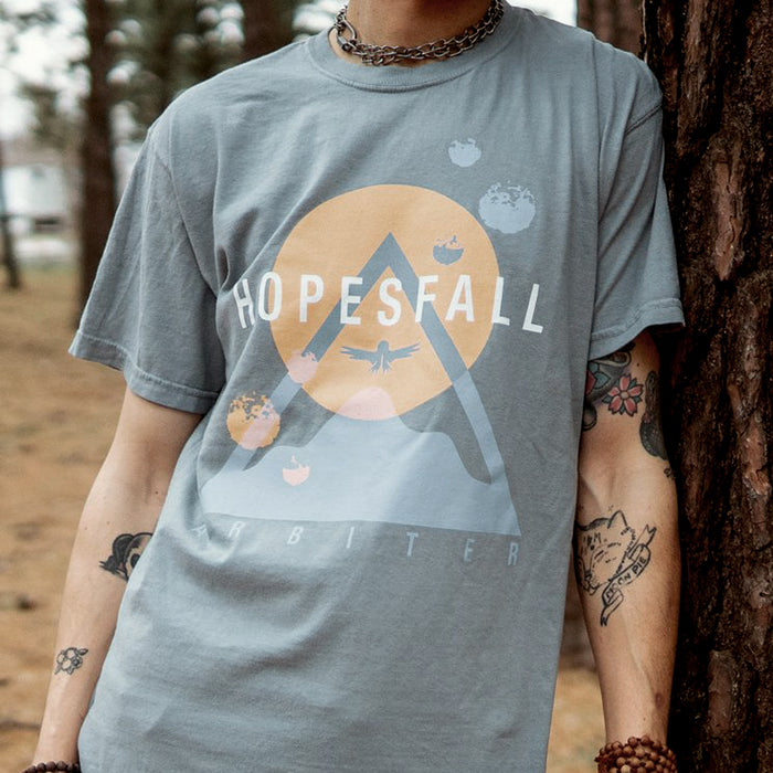 Grey short sleeve shirt with "HOPESFALL" written in white lettering across the chest. Behind that is an orange sun and a blue triangle making the shape of a mountain. There is a bird drawn over the sun in the same blue used for the mountain. Below the mountain is blue font that says "ARBITER". An individual is modeling the shirt, standing up against a tree.