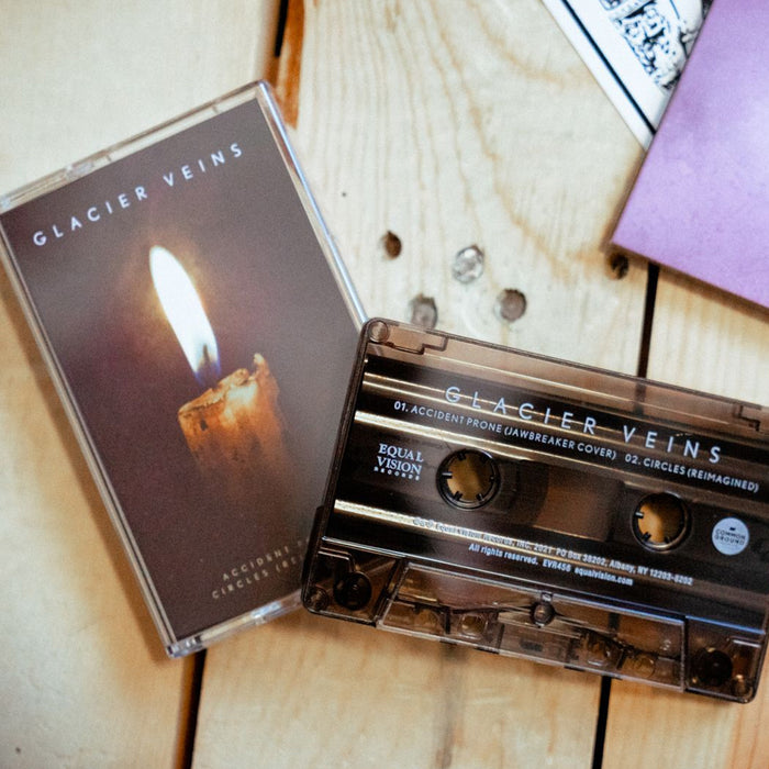 Cassette case with cassette on right hand side. Cassette case is a close up photo of a candle stick with a single flame. The top of the case says GLACIER VEINS in white lettering. On the side of the case, it says GLACIER VEINS in the same design. Cassette to the right of the case is black with a smokey tint. GLACIER VEINS written on top in white. Both are on top of light brown wooden planks.