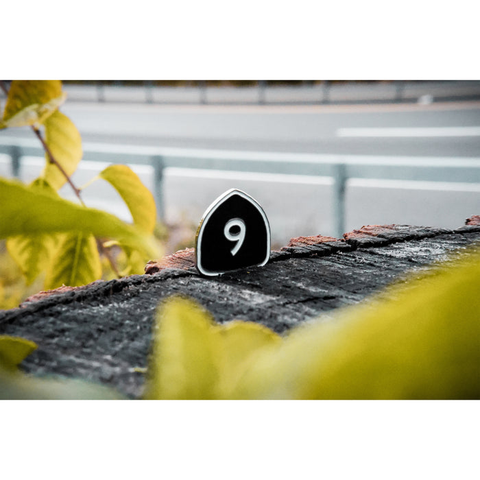 A small black sign with the number 9 stands on a weathered log among green and yellow leaves, evoking the cover of Saves The Day's Album 9, with a blurred road and guardrail in the background.