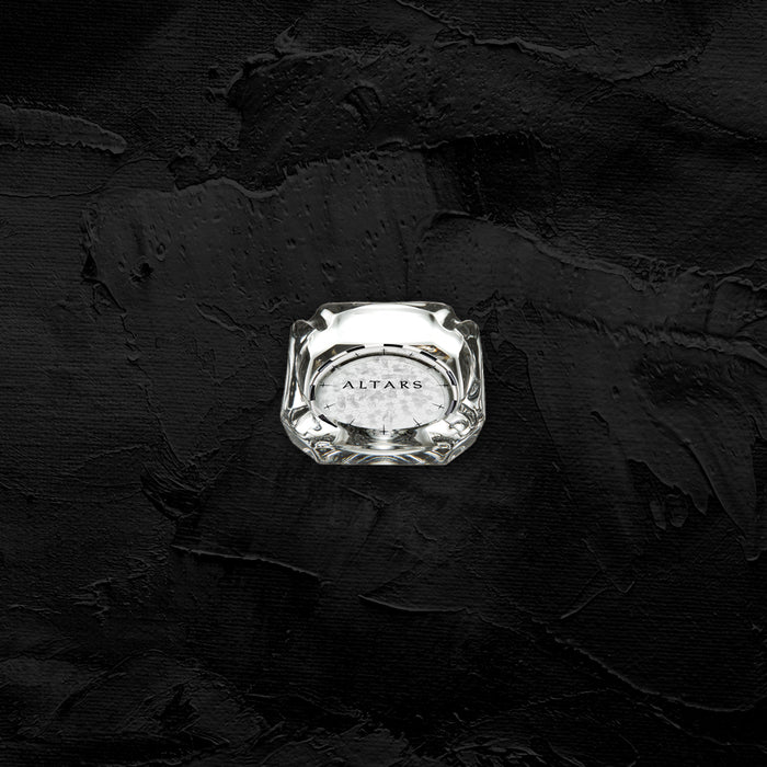 A clear glass perfume bottle labeled Sadistik sits centered on a textured, matte black surface. The minimalistic design highlights the bottle's allure and presence against the dark background.