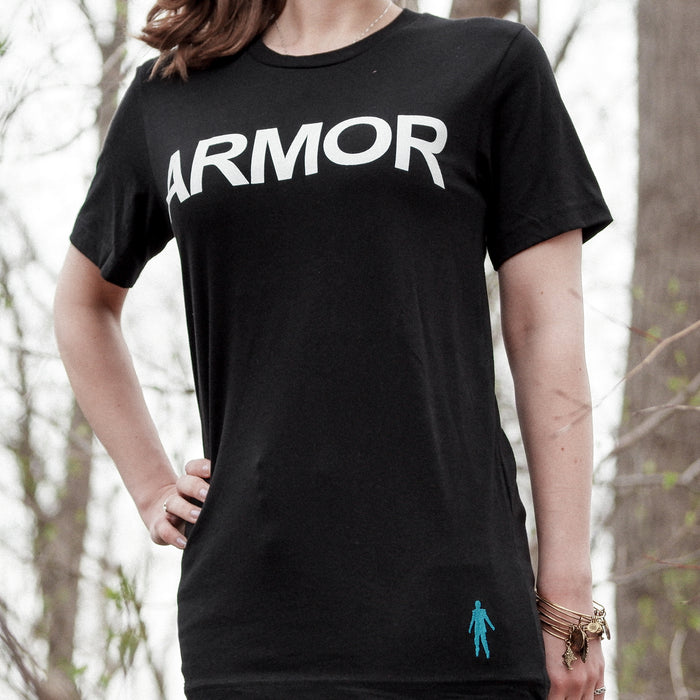 Black short sleeve shirt with ARMOR written on the chest in large white font. In the bottom right is a small threaded figure. An individual is wearing the shirt surrounded by trees.