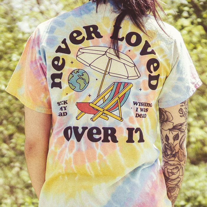 Back of multi-colored tie dye short sleeve. Beach chair and umbrella design in the center, with globe drawn in to the upper left. NEVER LOVED written above the beach chair, and OVER IT written below. To the left there is text that says STUCK IN MY HEAD, and to the right there is text that says WISHING I WAS DEAD. There is a model wearing the shirt, standing in front of greenery.