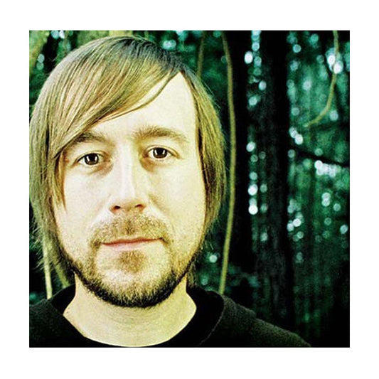 A man with straight, light brown hair and a beard stands outdoors before a forest background, channeling a Pop Rock vibe reminiscent of The Color Fred, as he looks at the camera with a neutral expression.