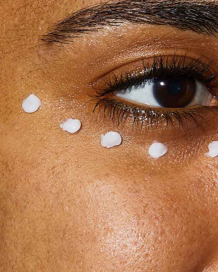 CeraVe Skin Renewing Eye Cream dots applied under a brown eye on medium skin tone, close-up on warm beige skin background.