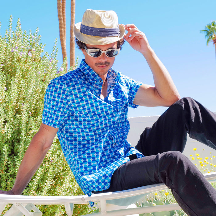 Man wearing a blue patterned shirt, sunglasses, and a straw hat sitting outdoors.