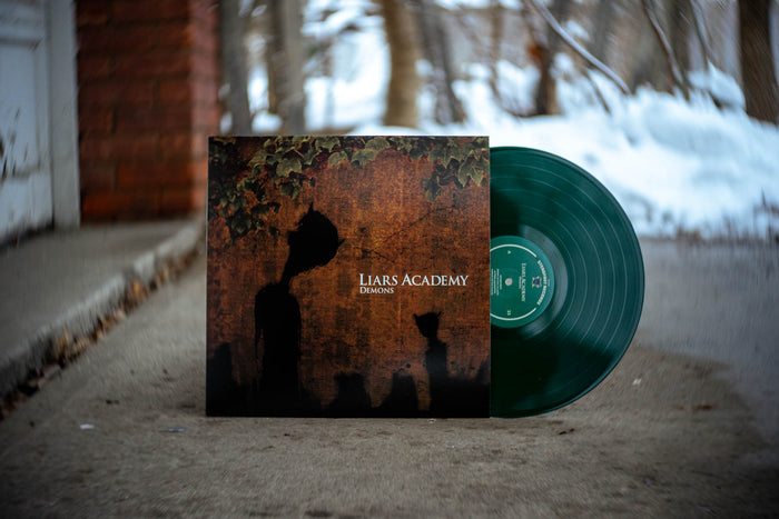 A green vinyl record and its cover, labeled Liars Academy, stand upright outdoors on concrete with snow and woods behind. The cover shows dark silhouettes and foliage, capturing a moody Baltimore-inspired scene.