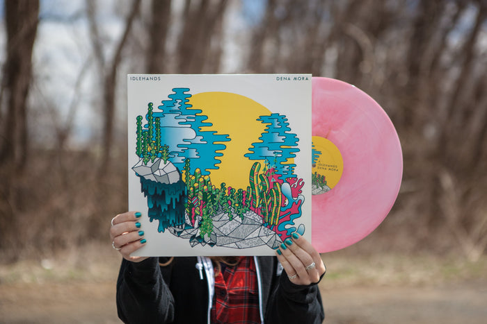 A person with teal-painted nails displays an IDLEHANDS Dena Mora vinyl, showcasing its vibrant cover art with abstract plants and a yellow sun. Limited pressing LP available and ready to ship.