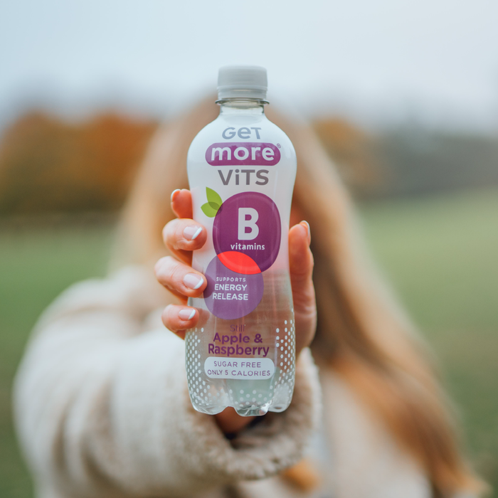 Hand holding a Get More Vits B Vitamins Still Apple & Raspberry 500ml bottle, labelled "Sugar Free Only 5 Calories", blurred outdoor background.
