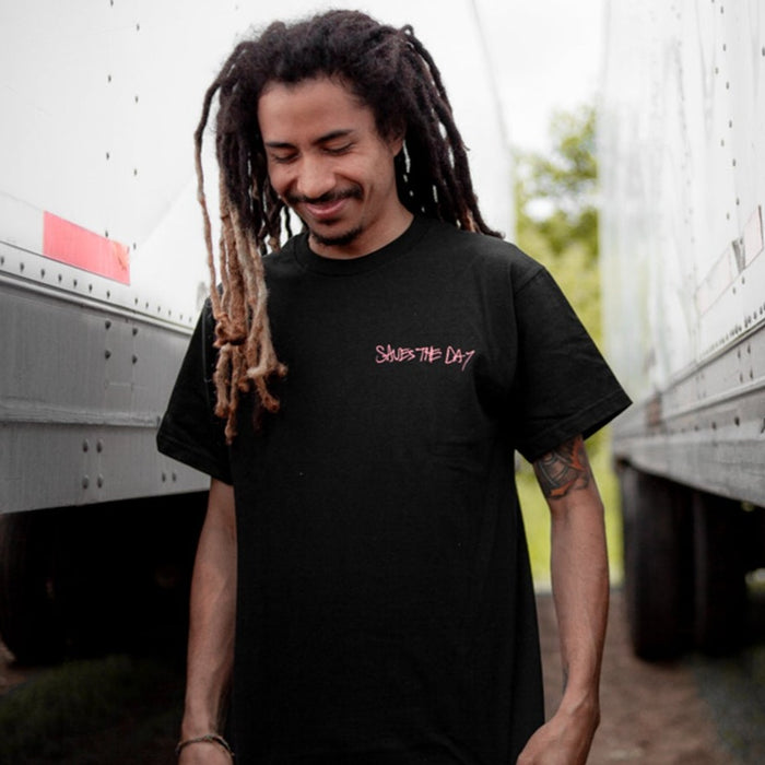 A man with long dreadlocks and a beard smiles while standing between two trucks, wearing a black Saves The Day t-shirt with the band name written in pink on the front.