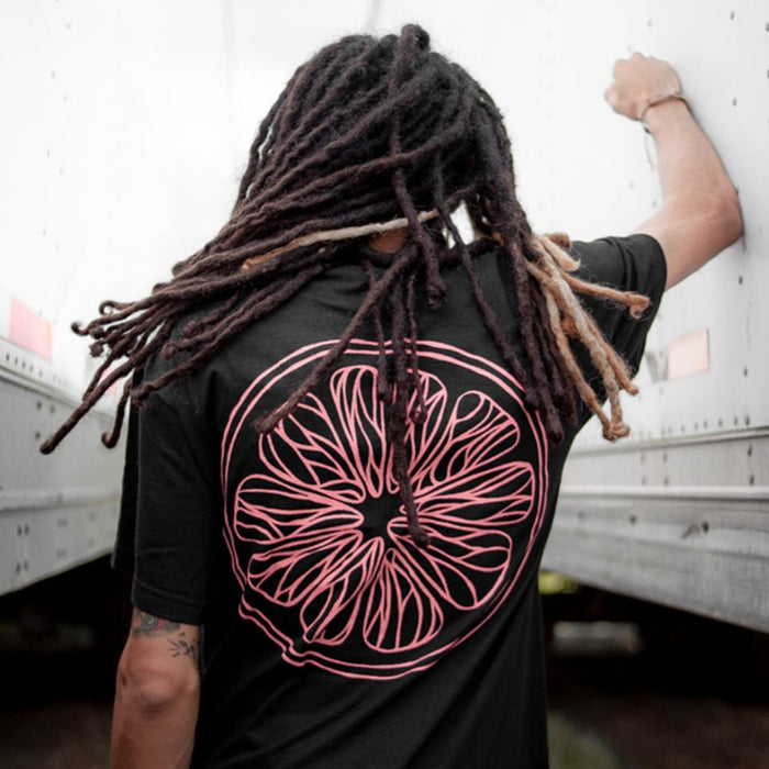 A person with long dreadlocks, seen from behind, wears a black Saves The Day T-Shirt with a large pink abstract citrus slice design while standing between two white vehicles, one arm resting on the side.