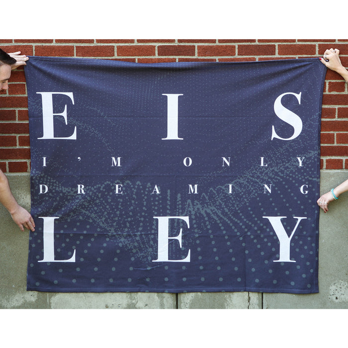Two people hold a cozy navy blue Eisley blanket with large white “EISLEY” letters and smaller “I’m only dreaming” text, set against a brick wall background.