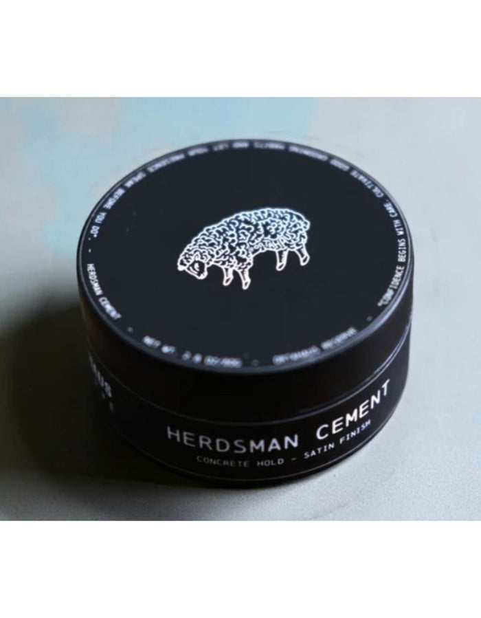 A black round tin of Herdsman Cement hair product with a white sheep logo and white text, photographed on a light gray surface.