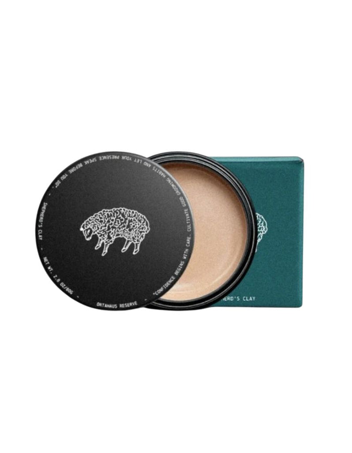 A round makeup compact with its lid off, showing a light tan pressed powder and a dark teal rectangular box behind it. The lid has a white sheep logo and text, and the box reads 'SHEPHERD'S CLAY'.