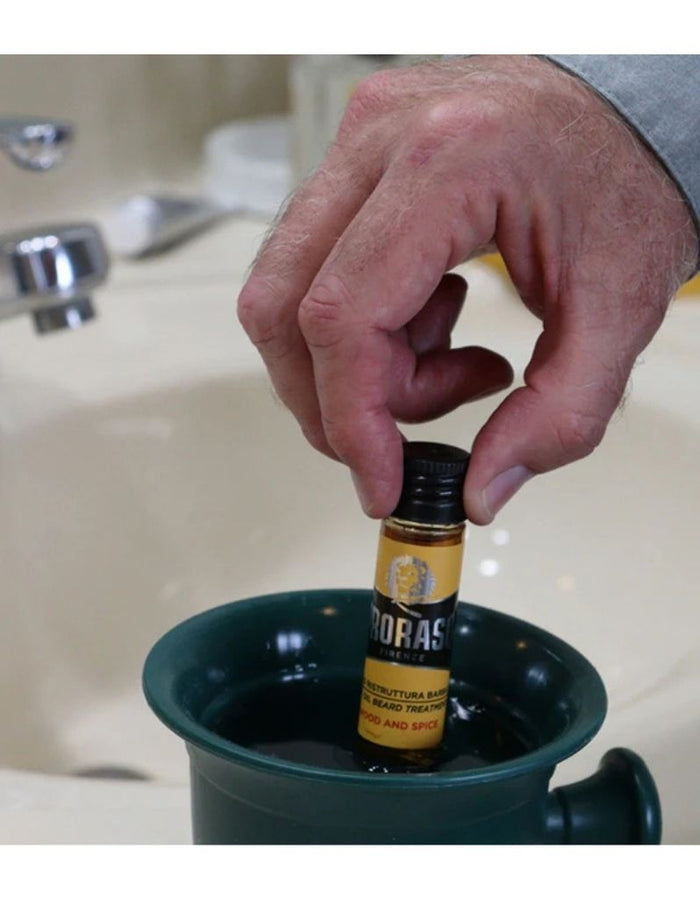 Close-up of a hand holding a small bottle of Proraso beard oil over a green bowl, with water in the background, demonstrating application process