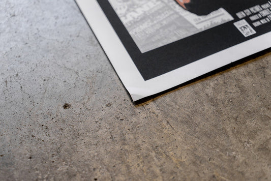 A close-up of a slightly damaged Bane 18x24 poster with a black border on gray concrete. The bottom corner is lifted, revealing text and a QR code, with no refunds faintly visible.