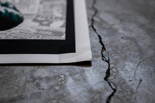 A close-up of the corner of a damaged 18x24 Bane poster lying on a cracked concrete floor, showing newspaper-like text and a dark border.