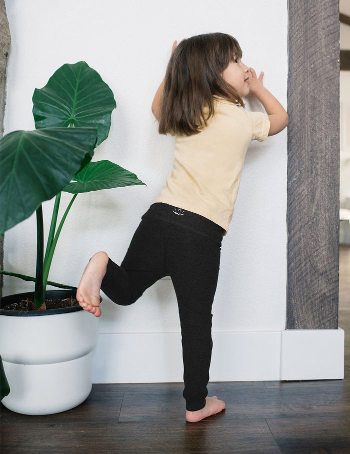 Toddler in black marled Spacedye Toddler Legging leans against a white wall next to a large potted plant, facing away.