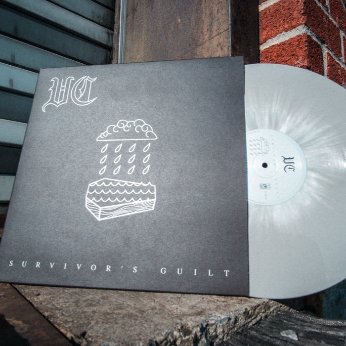 A limited edition grey-with-white-splatter vinyl by Vinnie Caruana partially slides from its sleeve, which shows a line drawing of a cake with rain and clouds. “VC” appears at the top and “SURVIVOR’S GUILT” at the bottom.