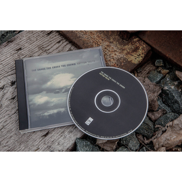 A CD case and disc for Cotton Teeth by The Snake The Cross The Crown rest on gravel and rusted metal; the cloudy sky cover hints at its atmospheric acoustic rock sound, while the disc features black with white text. Brand: The Snake The Cross The Crown.