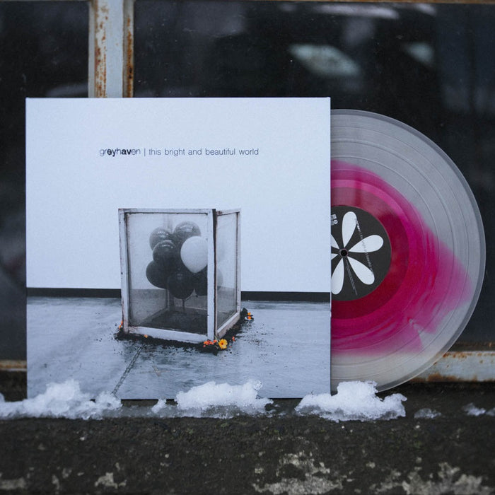 A limited edition Greyhaven vinyl with a milky clear and apple red spiral design is partially out of its cover, which shows black and white balloons in a glass box, with snow on the ground in front.