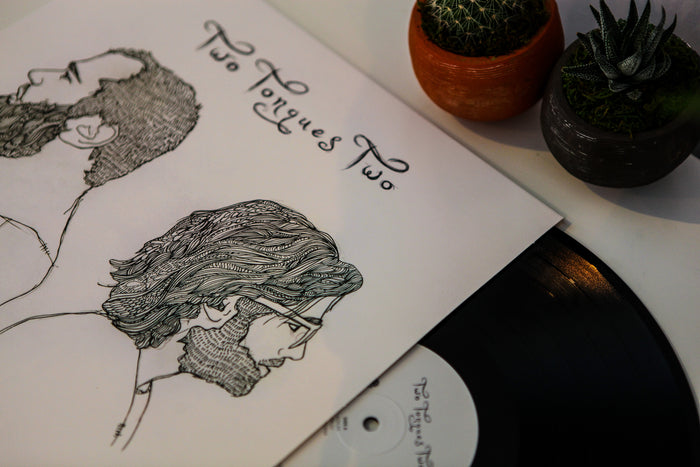 A limited edition black vinyl record by Two Tongues, partly out of its sleeve—which is illustrated with line drawings of two bearded men in profile and labeled Two Tongues LP—is displayed near two small potted succulents on a white surface.