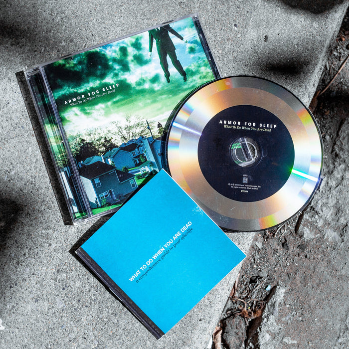 Square CD with white text that says ARMOR FOR SLEEP in blue and green tinted clouds. Below that there is smaller text that says WHAT TO DO WHEN YOU ARE DEAD. The album art is an image of a neighborhood of houses. In the top right there is a body of a man wearing a suit, but his head is not in the frame. On top of the cover is a reflective CD with a black inner circle. Inside the inner circle there is white text that says ARMOR FOR SLEEP. Both are laying on top of a concrete surface.