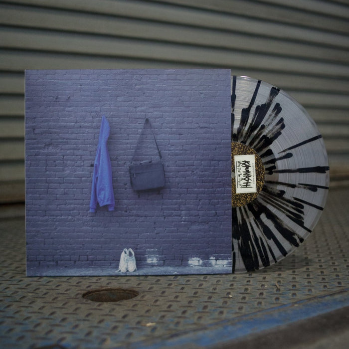 A photo of the vinyl and jacket propped up on a stand. The photo is taken on a metal warehouse gate. Clear vinyl with black splatter around it. Jacket of vinyl is a picture of a gray brick wall. A blue hoodie and black bag are hanging from the center of the brick wall. A pair of white sneakers are placed on the ground against the wall.