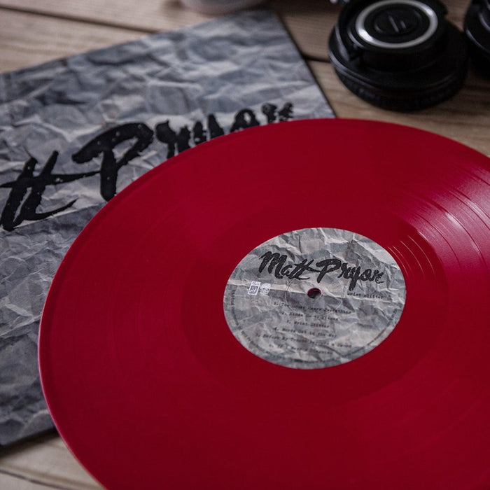 A red vinyl record by Matt Pryor, possibly a limited pressing LP, partially out of a crumpled sleeve with “Melt Prayer” text. Camera lenses rest nearby on a wooden surface.